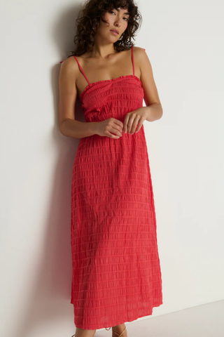 Model wearing the Nation LTD Madeline Tie Midi Dress in Flora, a coral pink textured midi dress featuring adjustable tie straps, shirred detailing, smocked voile fabric, a fitted bodice, and a flowing skirt.