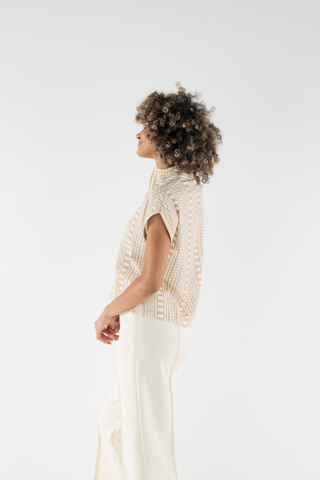 Woman wearing a beige knitted top and white pants on a light gray background