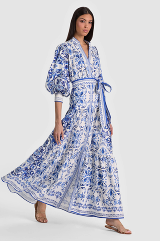Model wearing the Alice + Olivia Cora Maxi Shirt Dress in Solstice featuring a blue-and-white porcelain-inspired print, blouson sleeves, adjustable tie waist, tiered maxi skirt, and lightweight cotton voile fabrication paired with flat sandals