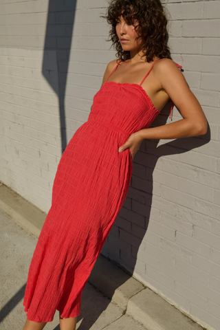 Model wearing the Nation LTD Madeline Tie Midi Dress in Flora, a coral pink textured midi dress featuring adjustable tie straps, shirred detailing, smocked voile fabric, a fitted bodice, and a flowing skirt.