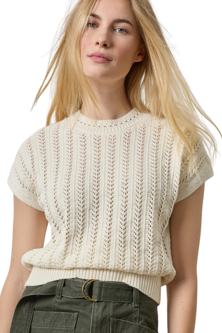 A woman wearing a cream-colored short-sleeve knit sweater with a textured vertical cable pattern, paired with dark olive green pants, against a plain white background.