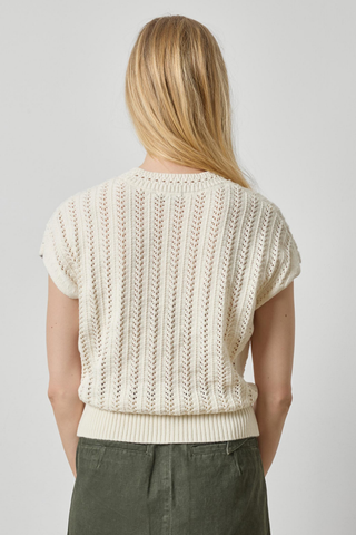 A woman viewed from the back wearing a cream-colored short-sleeve knit sweater with vertical textured stripes and olive green pants, set against a plain light gray studio background.