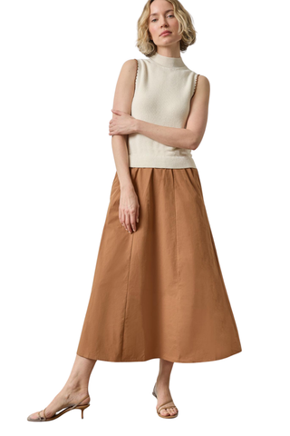 A woman modeling a sleeveless cream mock-neck knit top paired with a pleated brown midi skirt and beige heeled sandals, set against a plain white background.