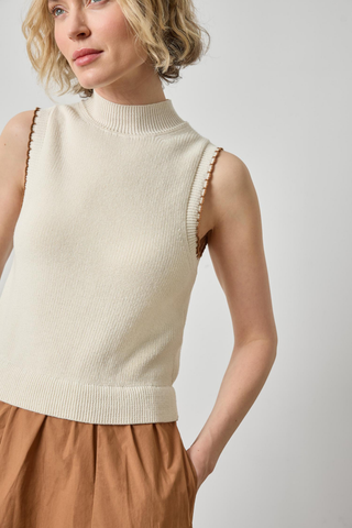 A woman wearing a sleeveless ribbed knit mock neck sweater vest in cream, paired with brown trousers, against a light gray studio background.