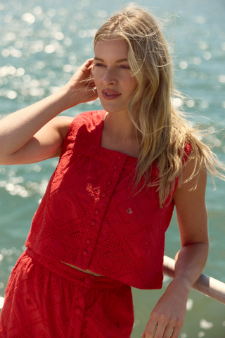 Model wearing the Olivia by Livro Nellie Top in Netta featuring vibrant coral eyelet embroidery, a sleeveless cropped silhouette, square neckline, covered button front detailing, and coordinating matching skirt styled with lace-up flat sandals