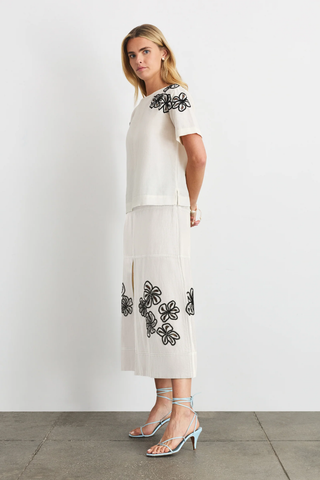 Woman wearing a white outfit with black floral embroidery against a plain background