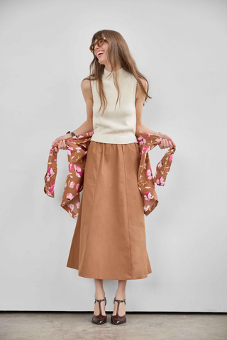 Woman wearing a cream sleeveless top and brown skirt with a floral scarf, standing against a plain background.