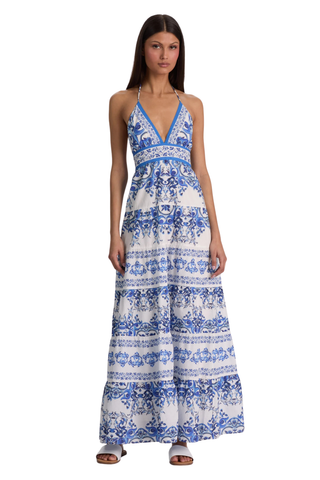 Model wearing the Alice + Olivia Karolina Maxi Dress in Solstice featuring a blue-and-white porcelain-inspired print, halter neckline, open back with braided tassel ties, smocked waist detail, and flowing tiered maxi silhouette paired with flat sandals