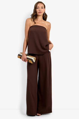 Woman wearing a brown strapless top and wide-leg pants holding a gold clutch against a white background