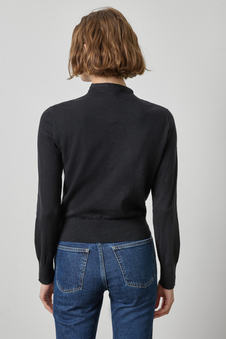 A woman viewed from the back wearing a black mock-neck long-sleeve knit top and blue jeans against a light gray studio background.
