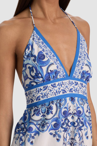 Model wearing the Alice + Olivia Karolina Maxi Dress in Solstice featuring a blue-and-white porcelain-inspired print, halter neckline, open back with braided tassel ties, smocked waist detail, and flowing tiered maxi silhouette paired with flat sandals