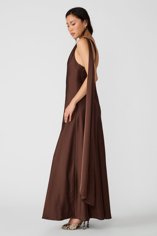 Steve Madden Aurorah Dress in Shaved Chocolate brown featuring a one-shoulder maxi silhouette, gold-tone hardware, and draped back scarf detail.