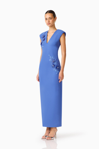 Model wearing the Elliatt Talitha Maxi Dress in Cornflower Blue featuring a plunging V-neckline, structured shoulders, floral appliqué detailing, and a slim column silhouette.