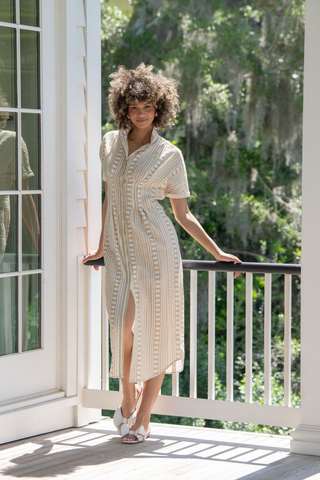 Woman in a patterned dress standing on a porch with greenery in the background