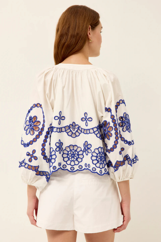 Model wearing the Marie Oliver Vara Top in Bluejay, an ivory cotton blouse featuring cobalt blue embroidered eyelet detailing, a tie neckline, puff sleeves, scalloped hem, and a relaxed silhouette.