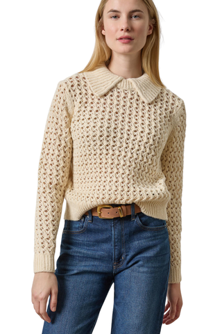 A woman wearing a cream-colored, textured knit sweater with a Peter Pan collar, tucked into blue jeans with a brown belt, against a plain white background.