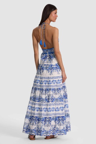 Model wearing the Alice + Olivia Karolina Maxi Dress in Solstice featuring a blue-and-white porcelain-inspired print, halter neckline, open back with braided tassel ties, smocked waist detail, and flowing tiered maxi silhouette paired with flat sandals