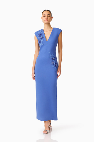 Model wearing the Elliatt Talitha Maxi Dress in Cornflower Blue featuring a plunging V-neckline, structured shoulders, floral appliqué detailing, and a slim column silhouette.