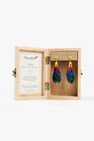 Model wearing the Brackish Leigh Statement Earring featuring handcrafted emerald green, royal blue, red, and yellow natural feathers with a 24K gold-plated stud and elongated teardrop silhouette.
