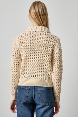 A woman viewed from the back wearing a cream-colored textured knit cardigan sweater and blue jeans against a light gray studio background.