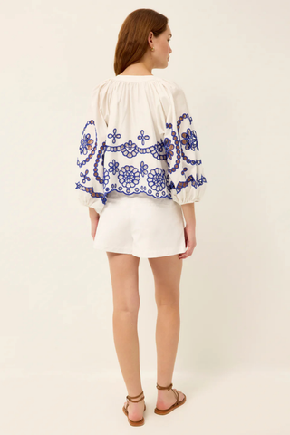 Model wearing the Marie Oliver Vara Top in Bluejay, an ivory cotton blouse featuring cobalt blue embroidered eyelet detailing, a tie neckline, puff sleeves, scalloped hem, and a relaxed silhouette.