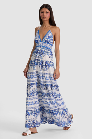 Model wearing the Alice + Olivia Karolina Maxi Dress in Solstice featuring a blue-and-white porcelain-inspired print, halter neckline, open back with braided tassel ties, smocked waist detail, and flowing tiered maxi silhouette paired with flat sandals