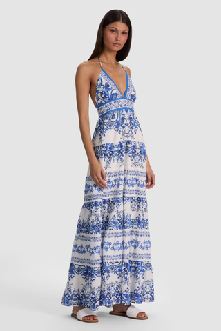 Model wearing the Alice + Olivia Karolina Maxi Dress in Solstice featuring a blue-and-white porcelain-inspired print, halter neckline, open back with braided tassel ties, smocked waist detail, and flowing tiered maxi silhouette paired with flat sandals