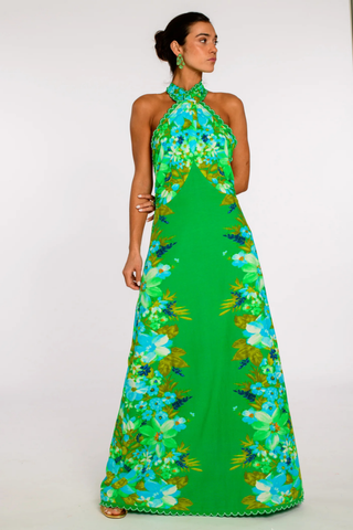 Model wearing the Elizabeth James the Label Aubrey Dress in Hula featuring a vibrant green tropical floral print, criss cross halter neckline, open smocked back, scallop trim detailing, and flowing maxi silhouette styled for a tropical resort setting