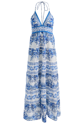 Model wearing the Alice + Olivia Karolina Maxi Dress in Solstice featuring a blue-and-white porcelain-inspired print, halter neckline, open back with braided tassel ties, smocked waist detail, and flowing tiered maxi silhouette paired with flat sandals