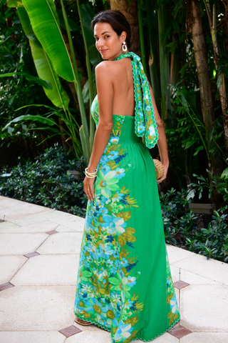 Model wearing the Elizabeth James the Label Aubrey Dress in Hula featuring a vibrant green tropical floral print, criss cross halter neckline, open smocked back, scallop trim detailing, and flowing maxi silhouette styled for a tropical resort setting