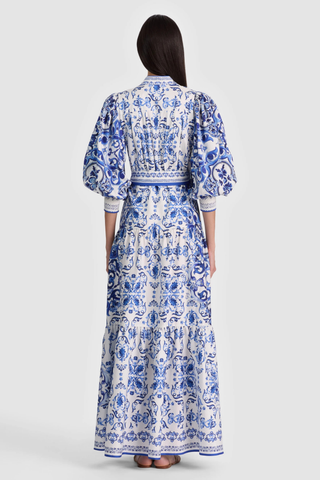 Model wearing the Alice + Olivia Cora Maxi Shirt Dress in Solstice featuring a blue-and-white porcelain-inspired print, blouson sleeves, adjustable tie waist, tiered maxi skirt, and lightweight cotton voile fabrication paired with flat sandals