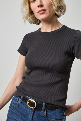A woman wearing a dark charcoal gray ribbed short-sleeve crewneck t-shirt tucked into blue jeans with a black belt featuring a gold buckle, photographed against a neutral light gray background.