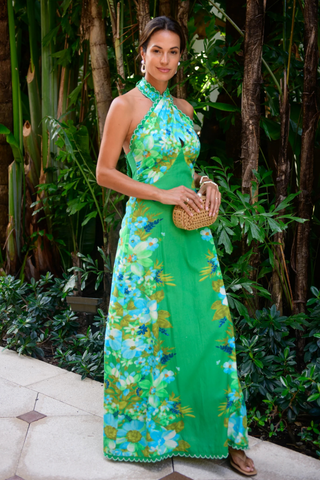 Model wearing the Elizabeth James the Label Aubrey Dress in Hula featuring a vibrant green tropical floral print, criss cross halter neckline, open smocked back, scallop trim detailing, and flowing maxi silhouette styled for a tropical resort setting
