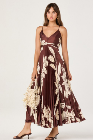 Model wearing the ASTR Clea Dress in Brown Floral featuring a gathered V-neck bodice, adjustable spaghetti straps, flowing pleated maxi skirt, and oversized cream floral print on a rich brown background.