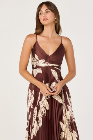 Model wearing the ASTR Clea Dress in Brown Floral featuring a gathered V-neck bodice, adjustable spaghetti straps, flowing pleated maxi skirt, and oversized cream floral print on a rich brown background.