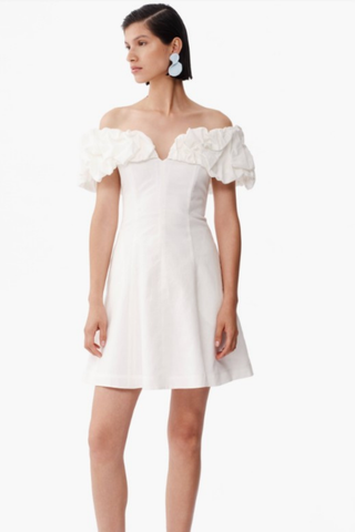 Model wearing De Loreta Cumaral Dress in White with off-the-shoulder ruffle sleeves, structured bodice, and fit-and-flare mini silhouette