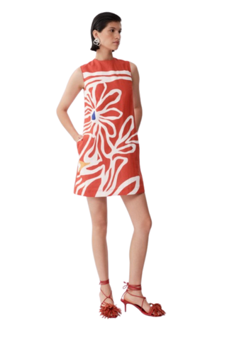 Model wearing De Loreta Mundo Dress in Miraflores with oversized ivory floral print, sleeveless shift silhouette, and coral red mini dress design